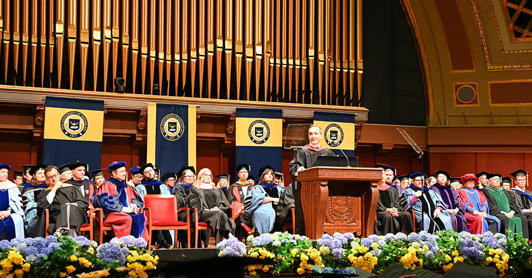 Graduation speaker Pedro Sancha at the podium at Hill Auditorium with faculty seated behind him