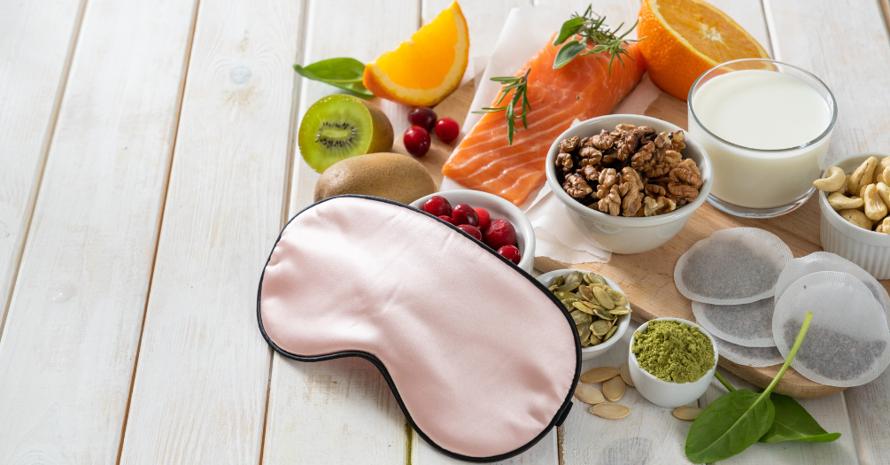 On a wooden table is a layout of different foods and a sleep mask. 
