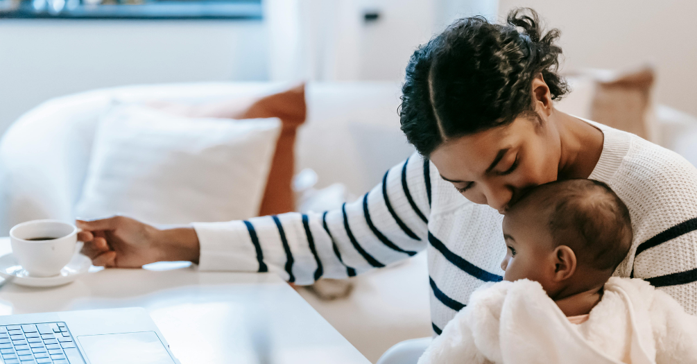 A mother kisses her child while working on her laptop.