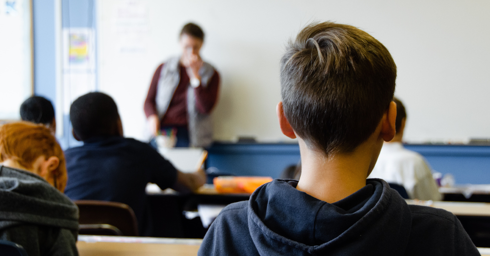 Students in classroom viewing presentation, shot from behind showing student in foreground and presenter at front of room