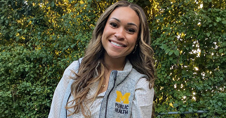 Juliana Taylor with a gray vest with Michigan Block M and words: Public Health standing in front of some green shrubs