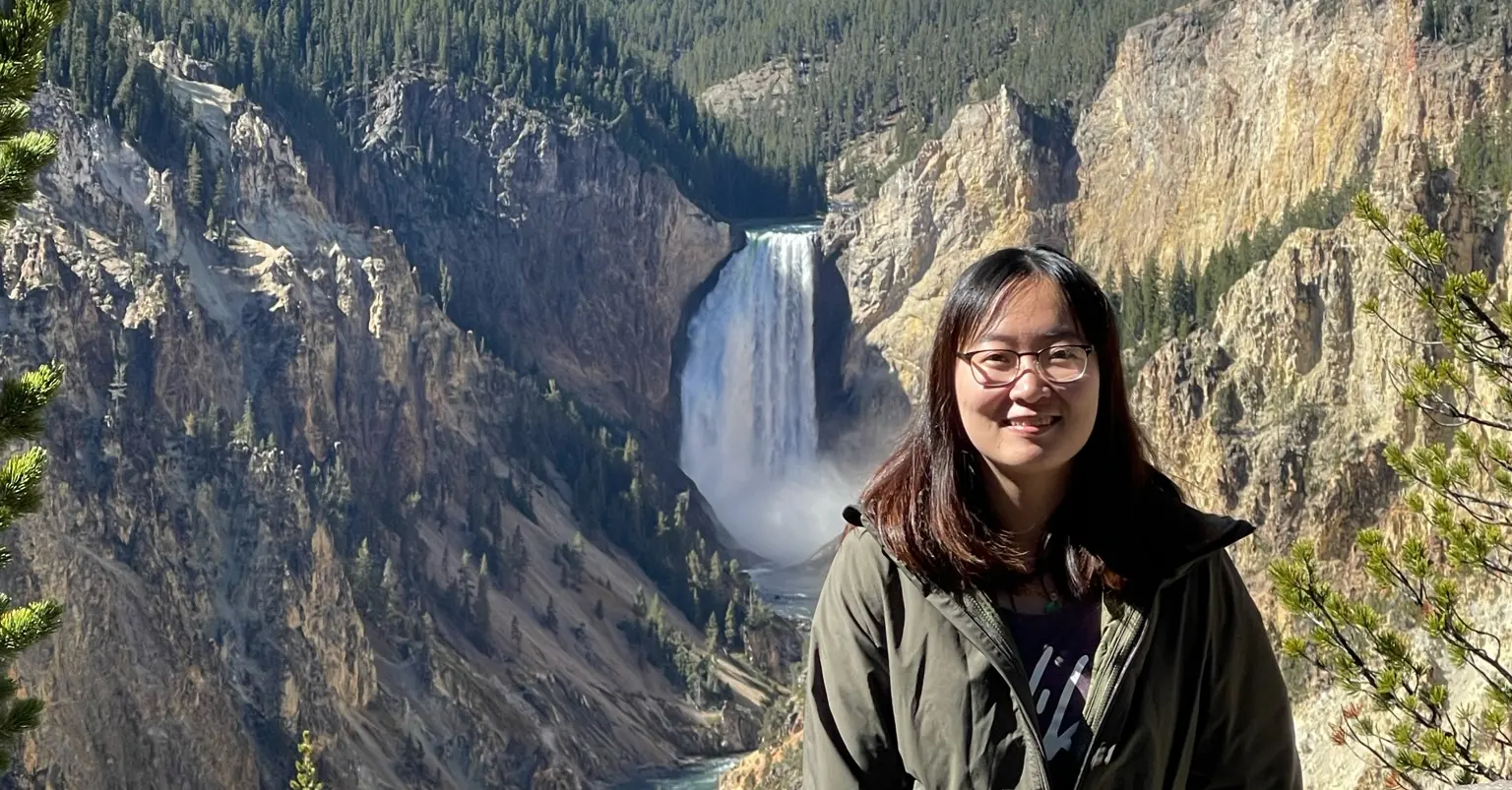 Liying Chen, wearing glasses and a green parka, with a waterfall in the background at Yosemite National Park